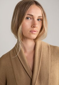 Beige gestrickter Wickelsweater mit breiten Revers, der über eine gerippte Textur und glatte Oberfläche verfügt. Das Kleidungsstück hat eine lockere Passform und minimalistische Details.