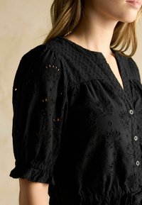 Schwarze Bluse mit kurzen, puffierten Ärmeln und Knopfleiste. Verfügt über bestickte Blumenmuster und einen strukturierten, atmungsaktiven Stoff.