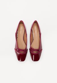 Ballerines en cuir verni bordeaux avec un bout rond, un nœud décoratif et un liseré élastique autour de l'ouverture. Texture lisse et brillante.