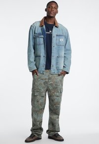Jeansjacke mit braunem Kragen, blauer Farbton, vier Taschen; Camouflage-Cargo-Hosen, lockere Passform, strukturiertes Material. Schwarze Sneaker.