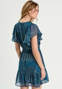 Robe turquoise avec des manches flottantes transparentes, taille smockée et ourlet à volants en étages. Présente un imprimé floral et géométrique avec des accents métalliques.