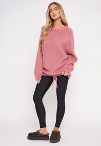Übergroßer pinker, flauschiger Pullover mit langen Ärmeln und Kordelzug am Saum, kombiniert mit schwarzen Leggings und braunen Slipschuhen.