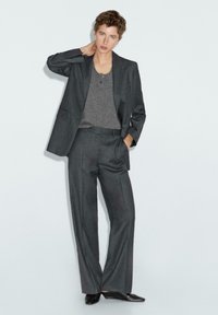 Dunkelgrauer Anzug mit einem taillierten Blazer, Bundfaltenhose und einem grauen T-Shirt darunter. Glattes Material mit einer subtilen Textur. Schwarze Schuhe.