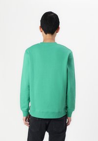 Sudadera de algodón verde con mangas largas, puños acanalados y cuello redondo. Llevada con pantalones vaqueros oscuros, vista desde la parte posterior contra un fondo blanco.
