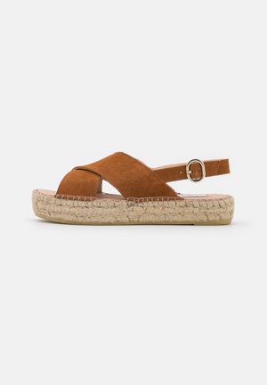 Brązowy sandał zamszowy z krzyżującymi się paskami, regulowany pasek wokół kostki z klamrą oraz pleciona platforma z juty na białym tle.