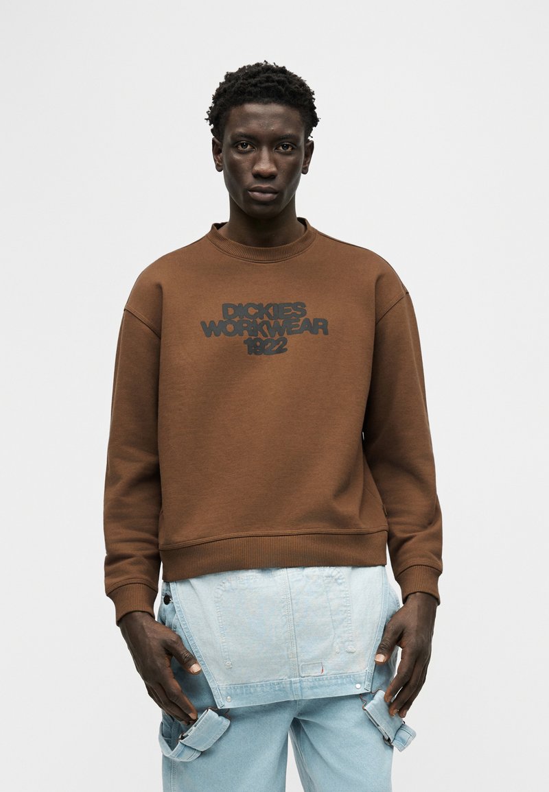 Young man wearing a brown "Dickies Workwear 1922" sweatshirt and light blue denim over pants against a plain background.