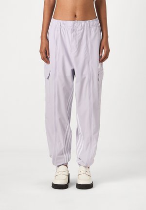 Person trägt hellviolette Cargo-Hose mit weißen Streifen an den Knöcheln und weiße Loafers mit Absatz, steht vor einem schlichten weißen Hintergrund.