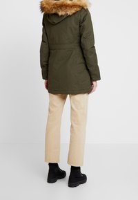 Parka verde olivo con capucha forrada de piel, hecha de una tela texturada. Presenta una silueta ajustada y se combina con pantalones beige y botas negras.