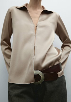 Personne portant une chemise beige en soie boutonnée partiellement ouverte à l'avant, un pantalon sombre et une large ceinture marron avec une grande boucle métallique.
