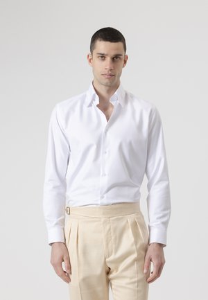 Joven con camisa blanca abotonada y pantalones plisados beige, de pie frente a un fondo liso gris claro.
