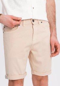 Shorts en denim beige présentant une coupe droite, des ourlets retroussés et des accents de boutons en métal noir, présentés portés avec une chemise blanche.