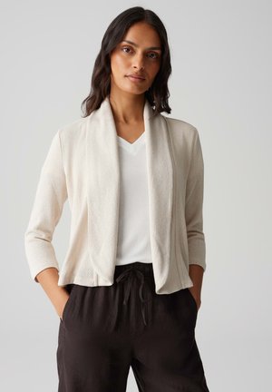 Lichtbeige cardigan met een gedrapeerde open voorkant, lange mouwen en een gestructureerde brei stof, gecombineerd met een witte v-hals top en zwarte broek.