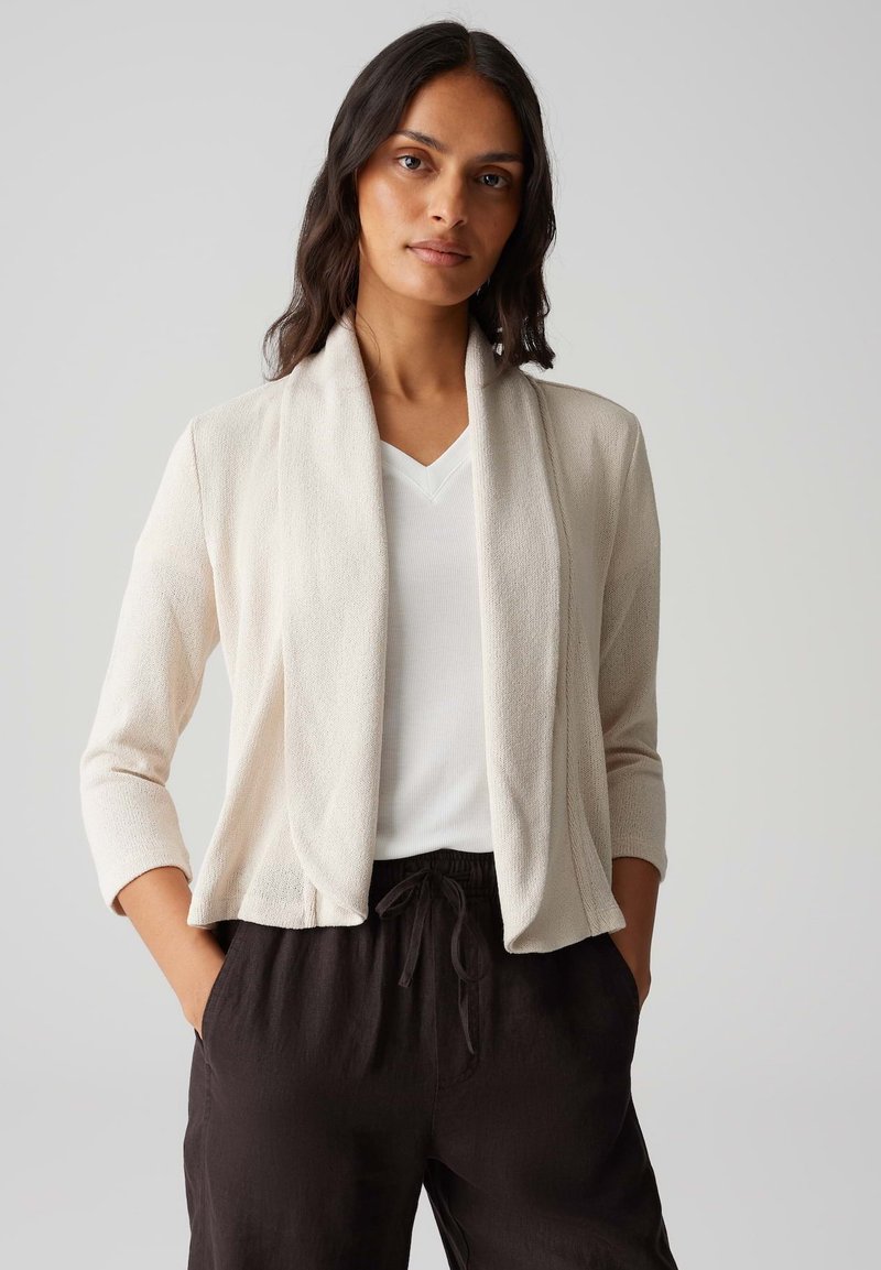 Helle beigefarbene Strickjacke mit offener Drapierung, langen Ärmeln und strukturiertem Strickstoff, kombiniert mit einem weißen V-Ausschnitt-Oberteil und schwarzen Hosen.