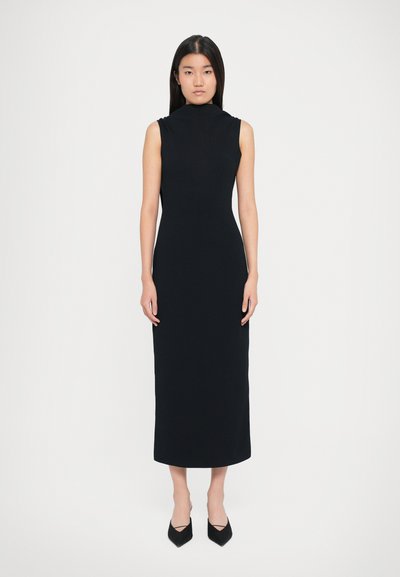 Schwarzes ärmelloses Midikleid mit hohem Kragen, figurnaher Silhouette und glatter Textur. Getragen mit schwarzen spitzen High Heels.