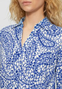Blauw en wit gestreepte blouse met een kraag en knopen, gemaakt van een lichtgewicht stof die abstracte vormen in verschillende tinten blauw toont.