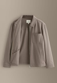 Veste taupe à fermeture éclair avec une poche poitrine et deux poches latérales, présentée ouverte sur un fond taupe assorti.