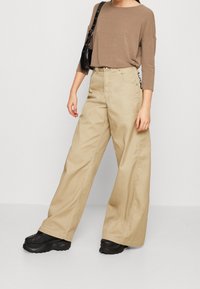 Beige weite Hose mit hohem Bund, aus Baumwollmaterial und sichtbaren Nähten. Kombiniert mit einem braunen Langarmoberteil und schwarzen Schuhen.