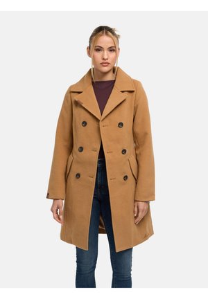 Vrouw draagt een dubbelgeknoopt camel coat over een donkere top en blauwe spijkerbroek, staande tegen een effen lichte achtergrond.