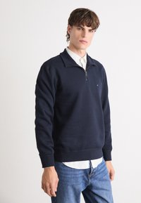 Navy blue zip-up sweatshirt made of soft fabric with a collared design, featuring a small logo on the chest and ribbed cuffs and hem.