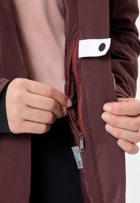 Handen die een bordeauxrode jaszak met een witte knoopsluiting openen, met zichtbare binnenvoering en label.