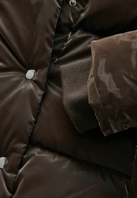 Hnědá péřová bunda s lesklým povrchem, vybavená stříbrnými druky a texturovanými pletenými manžetami. Hladká tkanina s prošívanými detaily.