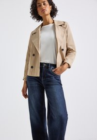 Veste beige croisée avec des boutons noirs, associée à un jean large bleu foncé et un t-shirt blanc. Tissu lisse et coupe ajustée.