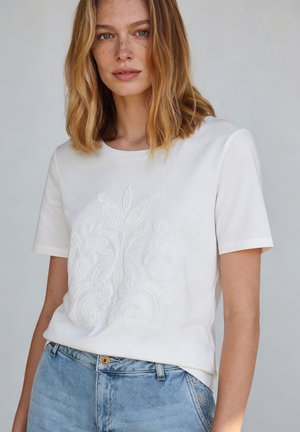 Junge Frau mit schulterlangem blondem Haar trägt ein weißes kurzärmeliges Shirt mit aufwendiger Blumenstickerei und hellblaue Jeans.
