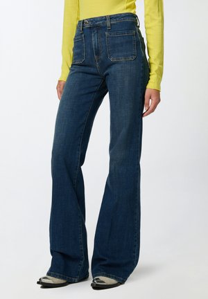 Frau trägt hoch taillierte, dunkelblaue Schlagjeans, gelbes Langarm-Shirt und spitz zulaufende Schuhe mit metallischen Spitzen, steht vor weißem Hintergrund.