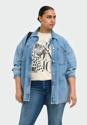 Femme en veste en jean et jean portant un t-shirt crème avec un grand graphisme léopard noir et blanc et un texte, regardant sur le côté.