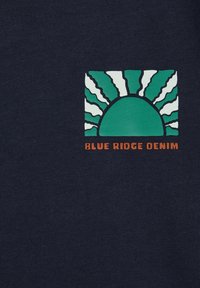 Jūras spēka audums ar grafiku, kurā attēlota zaļa saule, kas lec virs izliekta horizonta, akcentēta ar baltām stariem un tekstu "BLUE RIDGE DENIM" oranžā krāsā.
