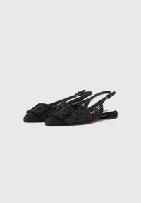 Chaussures à talon à lanière noires avec un design découpé floral complexe, talon plat et un doux détail en nœud rembourré sur l'orteil. Tissu texturé utilisé.