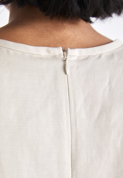 Light beige garment featuring a back zipper, smooth texture, and a simple rounded neckline. Seams are cleanly finished.