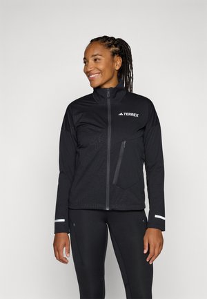 Svart Terrex-jacka med hög krage, zippered front, geometriska mönster och reflexdetaljer på ärmarna, tillsammans med svarta byxor.