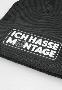 Black knit beanie featuring a rectangular patch with white text "ICH HASSE MONTAGE" and a graphic of a cat face. Textured exterior.