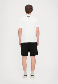 White short-sleeve t-shirt with a logo on the back, paired with black shorts and light sneakers, featuring pale socks.