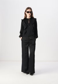 Blusa nera con dettagli arricciati sul davanti e chiusura con cravatta, abbinata a pantaloni neri a gamba larga. Modella con occhiali da sole neri e tacchi a punta.