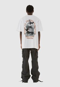 Hvid oversized T-shirt med grafik på ryggen, der viser et fuglebur og fugle, tekst i orange. Sammen med sorte cargo-bukser.