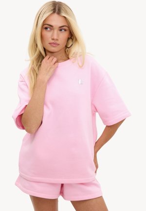 Femme blonde portant une chemise rose clair à manches courtes surdimensionnée et un short assorti, regardant sur le côté avec une main près du cou.