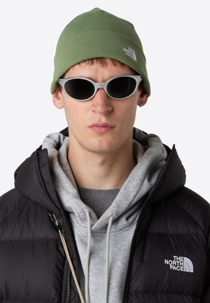 Joven con gafas de sol grises, gorro verde, sudadera gris y chaqueta de plumas negra de The North Face, mirando hacia adelante con una expresión neutral.