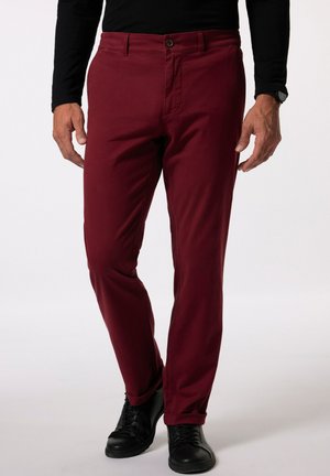 Homme portant un pantalon ajusté bordeaux, une chemise noire à manches longues, une montre noire et des chaussures noires à lacets, debout devant un fond uni.