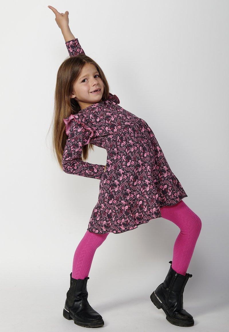 Vestido negro con estampado floral rosa, mangas rizadas, combinado con pantys rosa brillante y botas de tobillo negras. Cierre de cremallera ajustable en las botas.