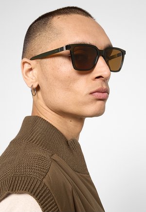 Persona con capelli corti che indossa occhiali da sole quadrati scuri, un piccolo orecchino a cerchio e un maglione di maglia marrone lavorato con collo alto.