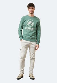 Sudadera verde con un logo circular y texto, combinada con pantalones cargo beige que tienen bolsillos laterales y zapatillas beige.