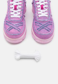 Paire de baskets en toile rose avec des lacets blancs et un motif bleu brodé, à côté d’un rouleau de lacets blancs sur une surface blanche.