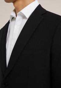 Svart skräddarsydd kavaj med snibbkrage, bröstficka och en slät yta. Bärs över en fräsch vit skjorta med knäppning.