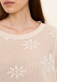 Žena v béžovém pleteném topu s květinovým vzorem a kulatým výstřihem, zachycena od krku po horní část hrudníku.