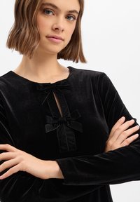 Schwarzes Samtoberteil mit rundem Ausschnitt, zwei kleinen Schleifen vorne und langen Ärmeln mit glatter Textur.