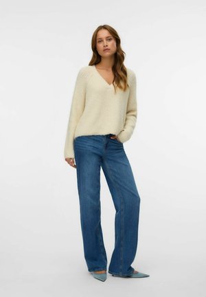 Beige gebreide trui met V-hals, gecombineerd met lichtblauwe wijde jeans. Het model draagt puntige blauwe schoenen, wat zachte texturen en een casual stijl benadrukt.