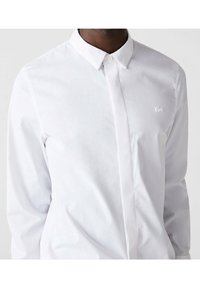Camisa blanca de manga larga en mezcla de algodón, con un collar clásico y un sutil bordado del logo en el pecho. Textura suave con un diseño ajustado.