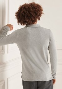 Langärmliges graues Poloshirt aus weichem Stoff, mit einem Kragen und einem kleinen Seitenschlitz am Saum. Glatte Textur mit minimalen Details.